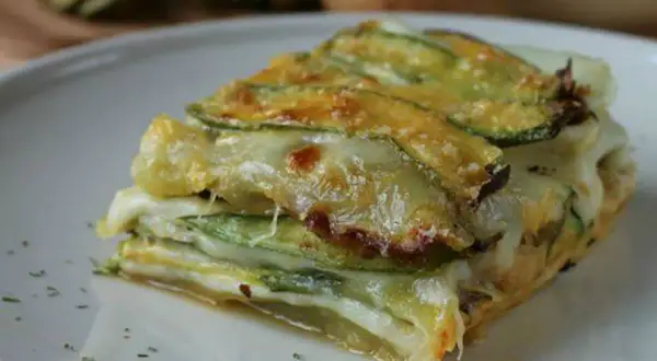 Una fetta di parmigiana di zucchine bianca, stratificata con formaggio filante e zucchine grigliate, servita su un piatto bianco.