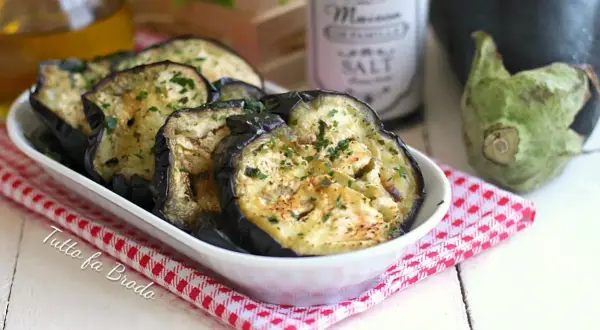 Fette di melanzane in padella, dorate e profumate con prezzemolo, servite in una ciotola bianca su una tovaglietta a quadretti rossi.