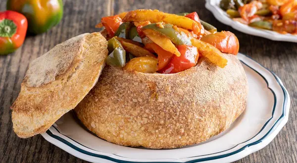 Un pane scavato ripieno di un saporito stufato di pipi e patate, con una fetta di pane accanto.