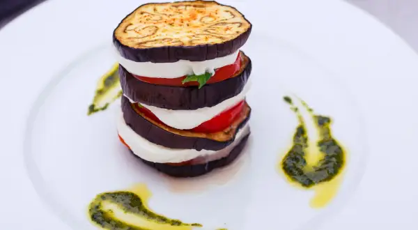 Parmigiana di melanzane grigliate a strati con mozzarella e pomodoro, guarnita con pesto e olio.