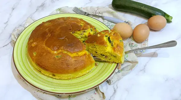 Torta salata zucchine e speck dorata su piatto verde, con ingredienti freschi a lato.