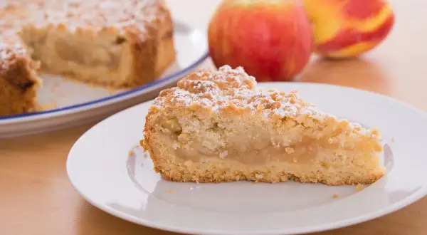 Una fetta di sbriciolata di mele, cosparsa di zucchero a velo, su un piatto bianco. Sullo sfondo, una torta intera e mele rosse.