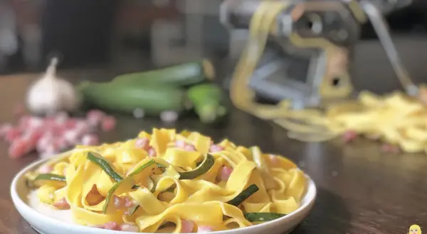 Piatto di pasta fresca con zucchine e pancetta, ingredienti freschi e macchina per la pasta sullo sfondo.