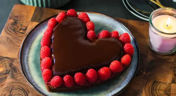 Deliziosa torta a forma di cuore ricoperta di cioccolato fuso e decorata con lamponi freschi, perfetta per un'occasione speciale.