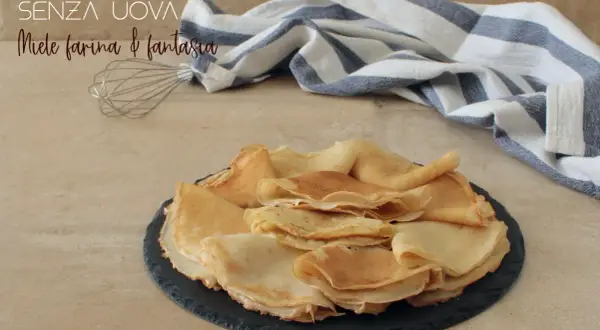 Crepes senza uova, dorate e soffici, impilate su un piatto scuro. Un battitore e un tovagliolo a righe completano la scena.