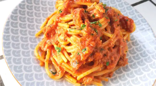 Un piatto di spaghetti al tonno, conditi con una ricca salsa rossa e guarniti con prezzemolo tritato, serviti su un piatto con motivo a squame.