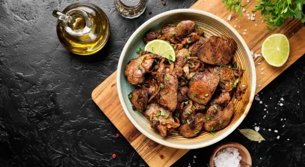 Piatto di fegatini di pollo saltati con cipolle, guarniti con prezzemolo fresco e spicchi di lime. Accanto, olio d'oliva, sale grosso e pepe.