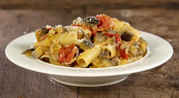 Piatto di pasta con melanzane e pomodorini, condita con formaggio grattugiato, su un tavolo di legno.