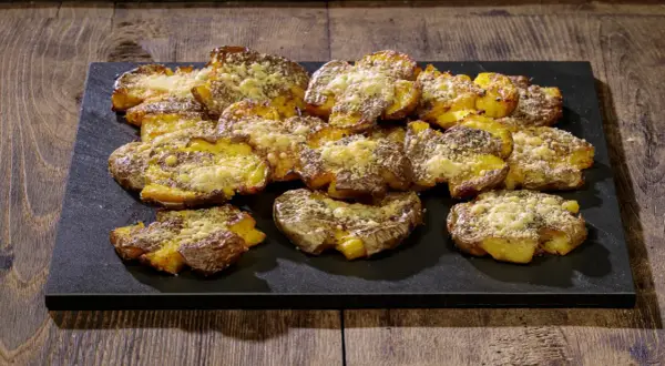 Patate schiacciate al forno, dorate e croccanti, cosparse di formaggio grattugiato e erbe aromatiche, servite su una pietra scura.