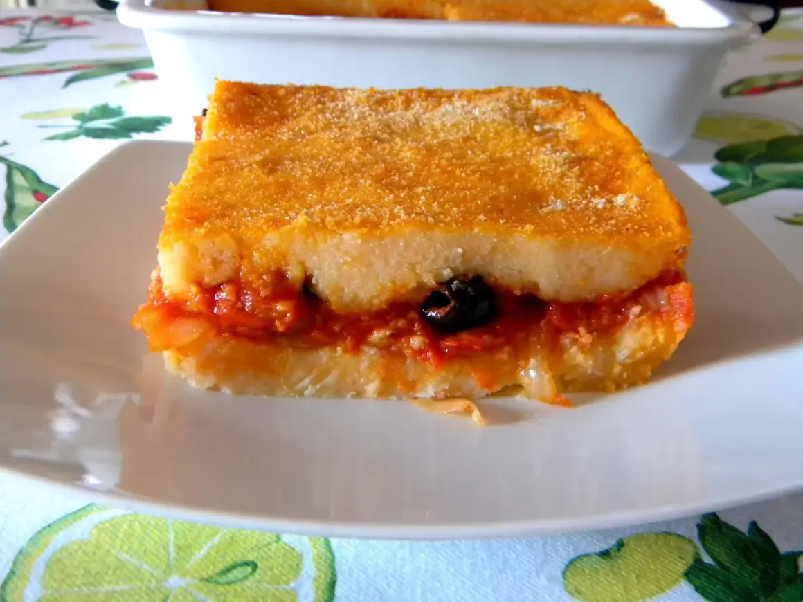 Una fetta di pitta di patate dorata e ripiena di sugo, cipolle e un'oliva nera, servita su un piatto bianco.