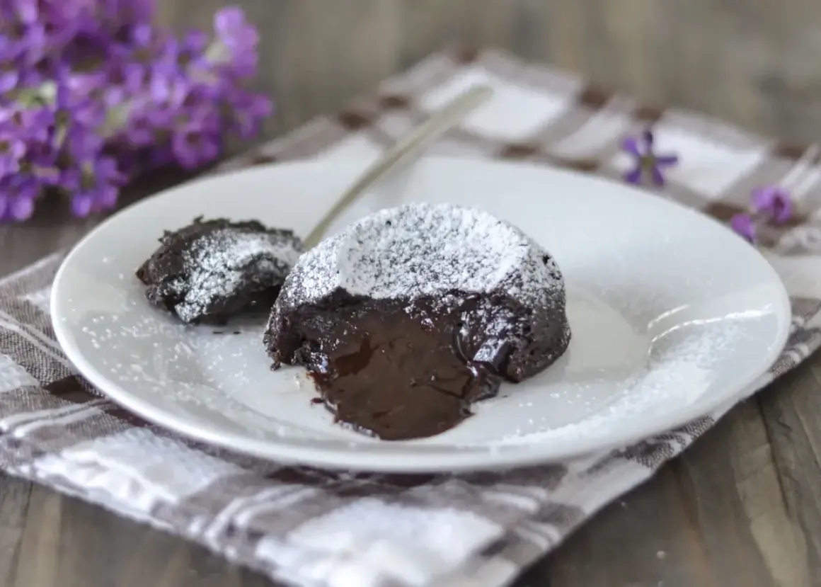 Un delizioso tortino al cioccolato con cuore fuso, spolverato di zucchero a velo, su un piatto bianco con fiori viola sullo sfondo.