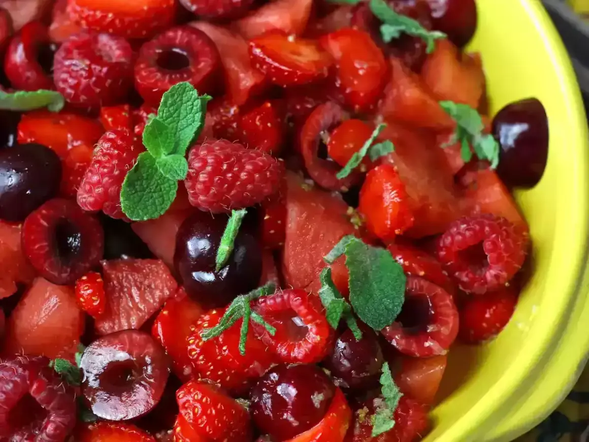 Macedonia di fragole, lamponi e ciliegie, guarnita con foglie di menta fresca, in una ciotola gialla.