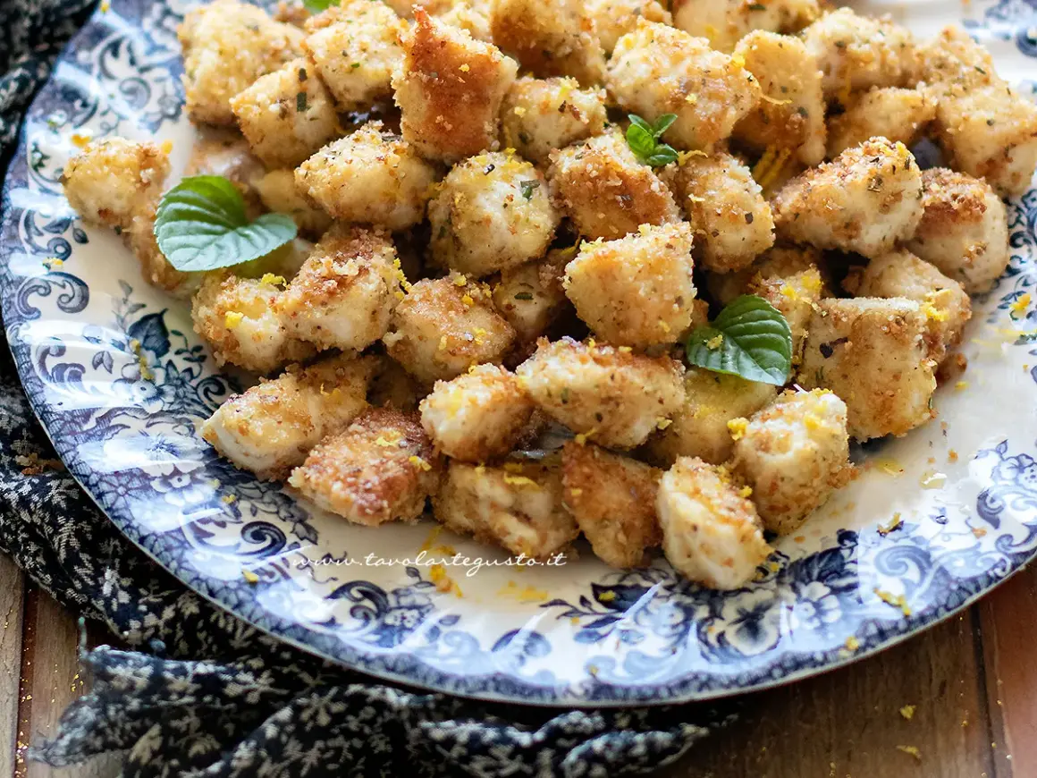 Bocconcini di pollo al forno dorati e croccanti, guarniti con scorza di limone e foglie di menta fresca su un piatto decorato.