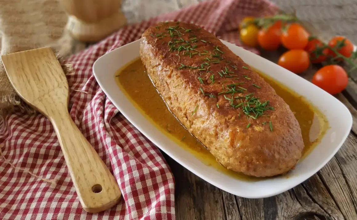 Polpettone dorato in padella, guarnito con erba cipollina, su un piatto bianco con salsa, accanto a pomodorini e una spatola di legno.