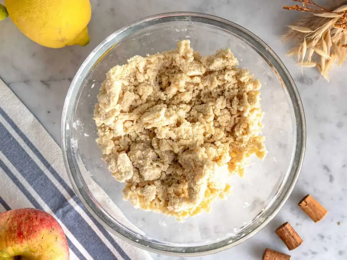 Impasto friabile per crumble di mele in una ciotola di vetro, con limone, mela e cannella sullo sfondo.
