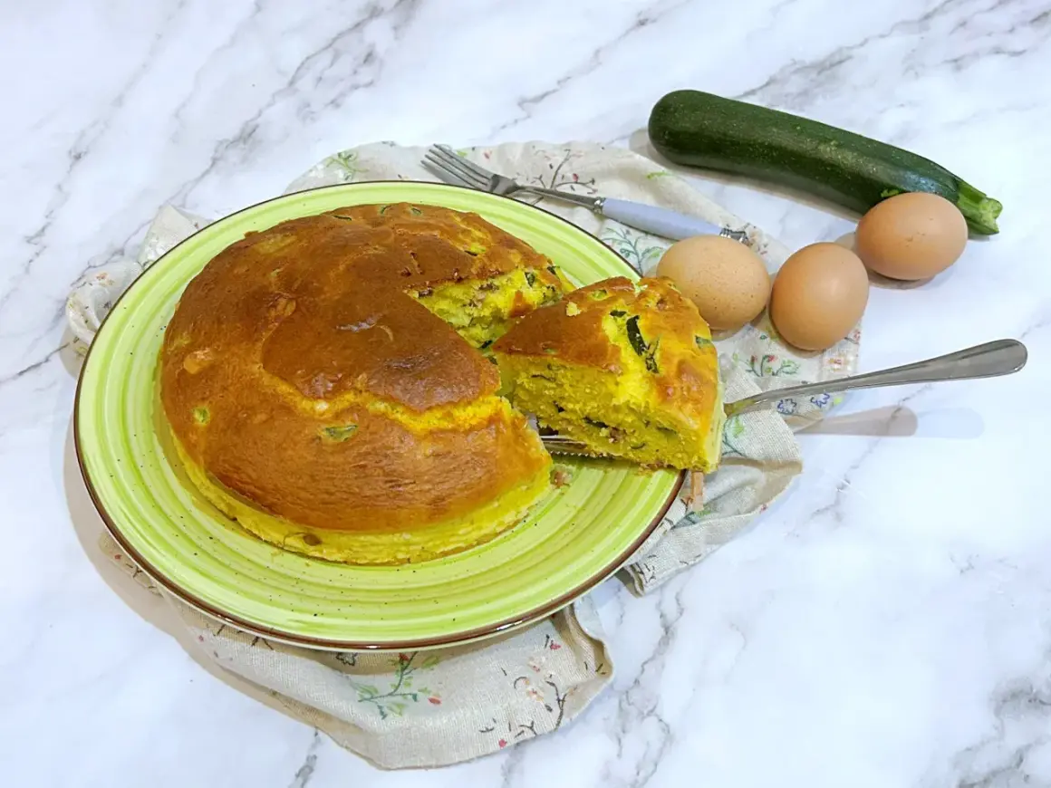 Torta salata zucchine e speck dorata su piatto verde, con ingredienti freschi a lato.
