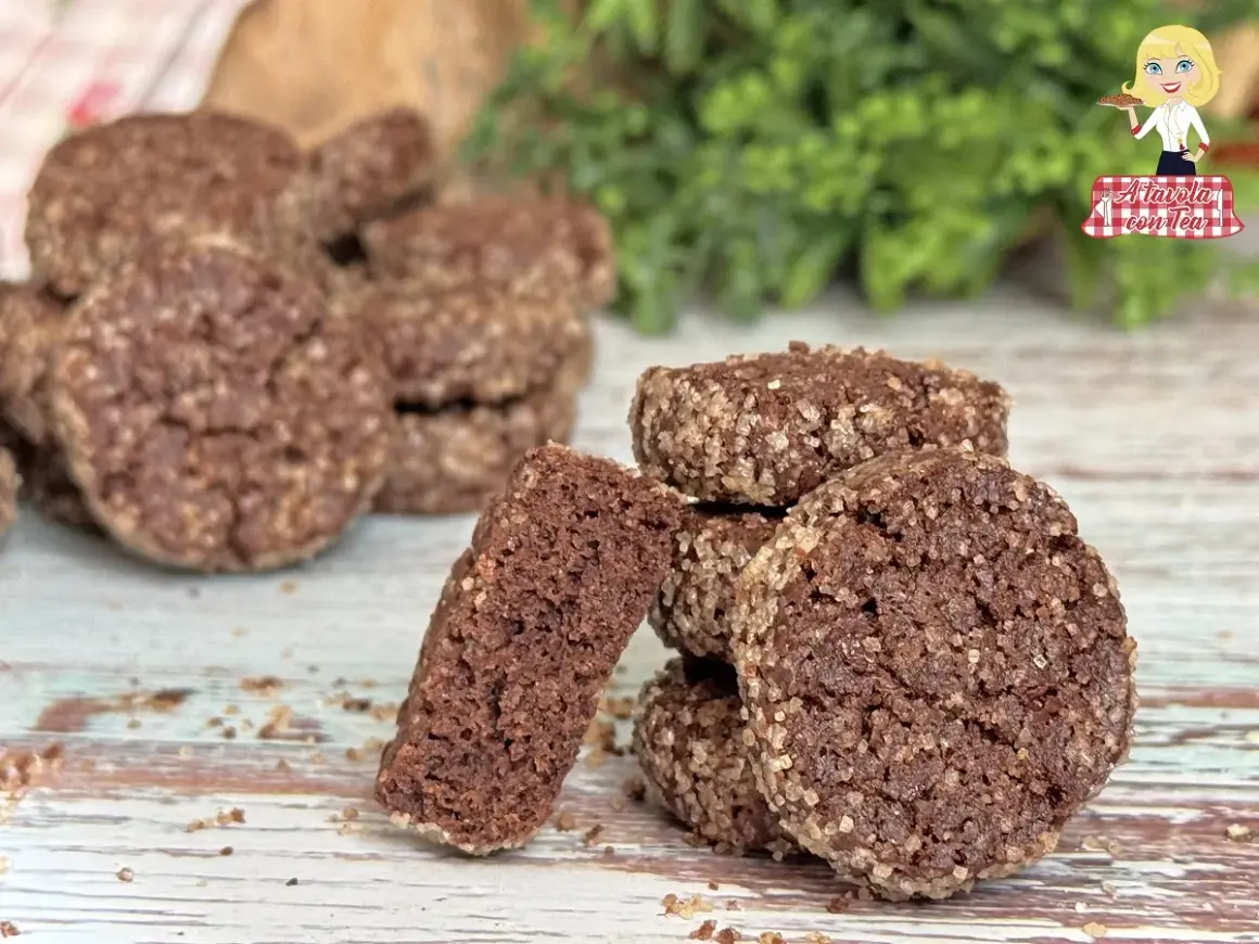 Biscotti al cioccolato rustici, ricoperti di zucchero grezzo, su un tavolo di legno. Uno spezzato rivela la consistenza friabile.