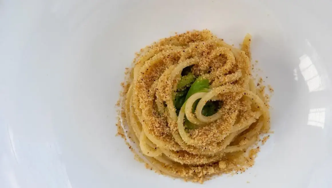 Un piatto di pasta alla carrettiera, spaghetti conditi con mollica tostata e verdure verdi, serviti su un piatto bianco.