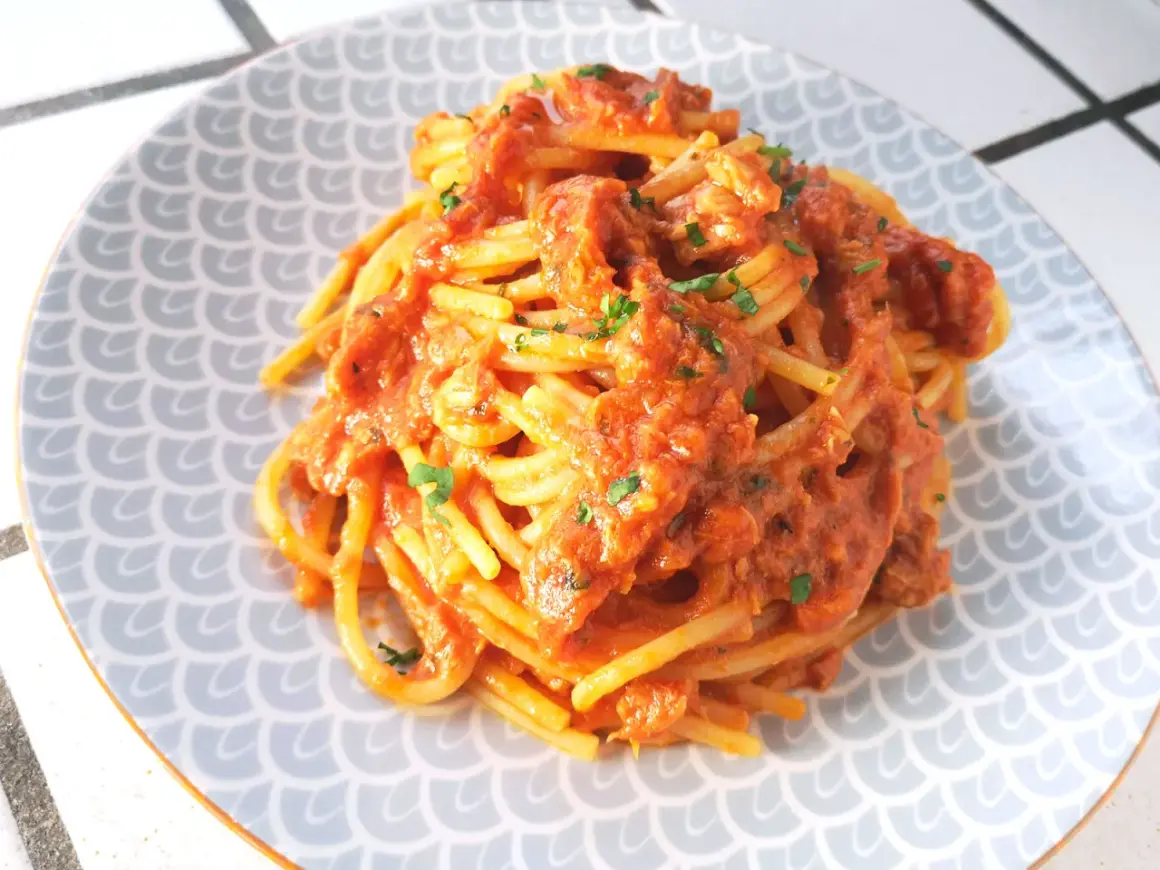 Un piatto di spaghetti al tonno, conditi con una ricca salsa rossa e guarniti con prezzemolo tritato, serviti su un piatto con motivo a squame.