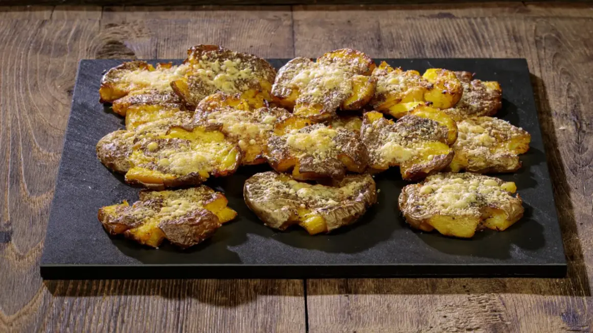 Patate schiacciate al forno, dorate e croccanti, cosparse di formaggio grattugiato e erbe aromatiche, servite su una pietra scura.
