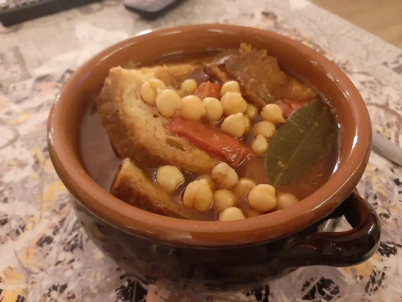 Minestra di ceci fumante in ciotola di terracotta, con pane raffermo, pomodoro e foglia di alloro.