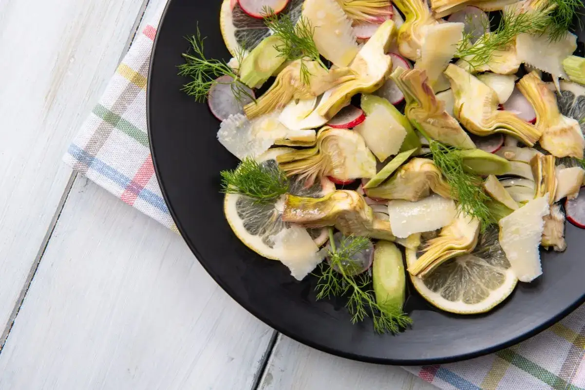 Deliziosa insalata di carciofi crudi con scaglie di parmigiano, spicchi di limone, ravanelli e finocchietto fresco su piatto nero.
