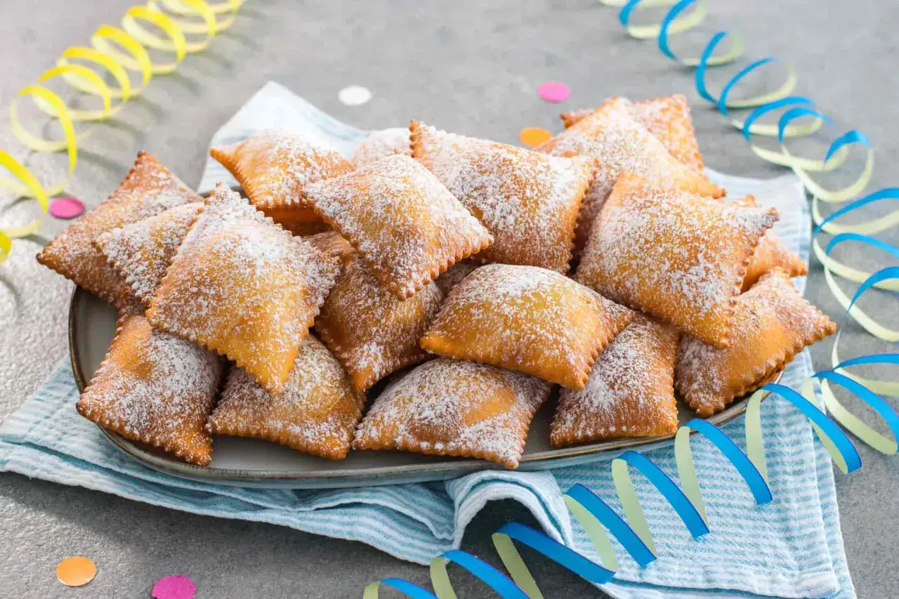 Bugie ricetta originale: dolcetti fritti a forma quadrata, cosparsi di zucchero a velo, su un piatto con festoni colorati.