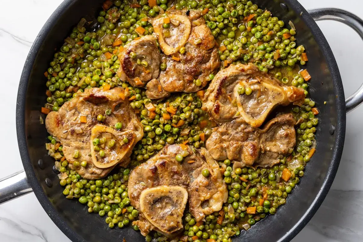 Quattro fette di ossobuco con piselli e carote in una padella. Un piatto saporito e colorato, pronto per essere gustato.