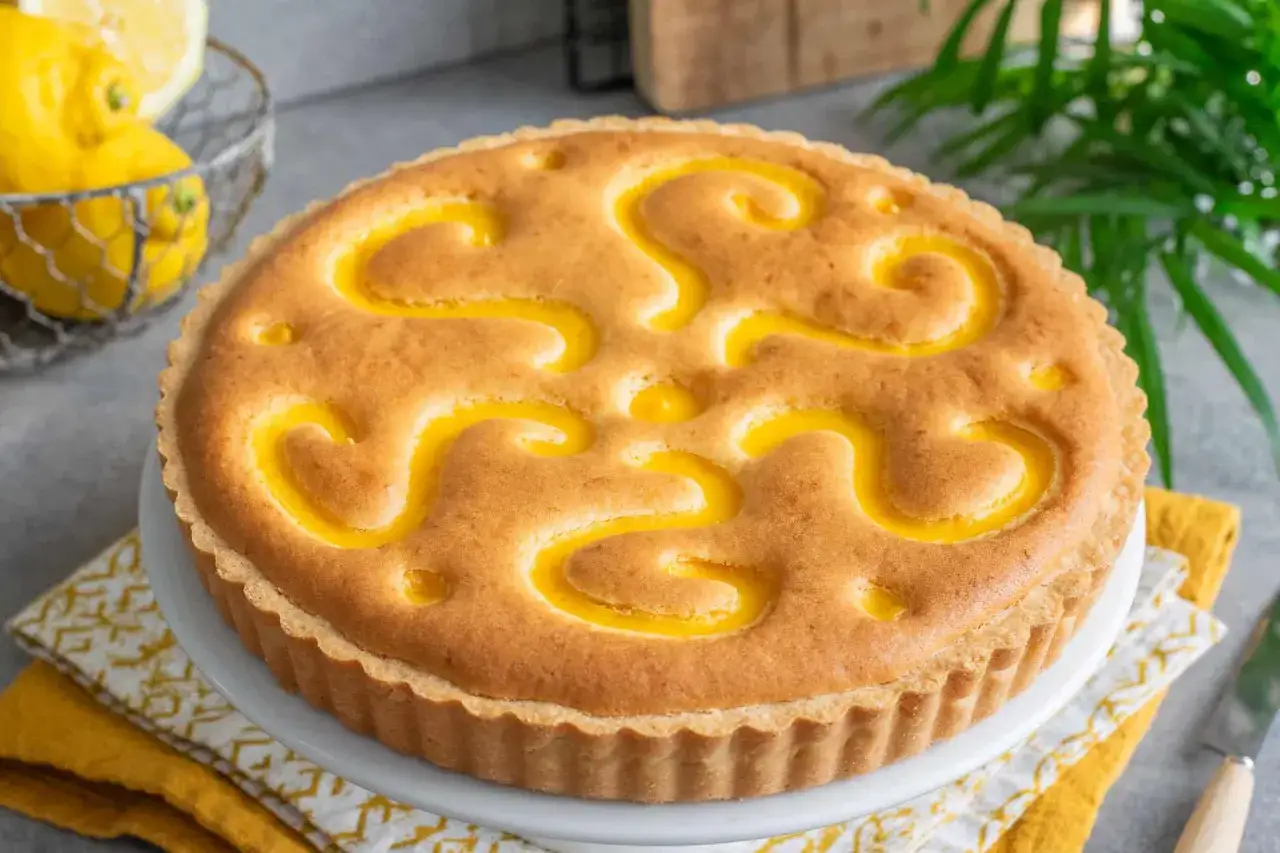 Crostata con crema pasticcera, decorata con un motivo a spirale giallo brillante, su un piedistallo bianco. Limoni freschi in un cestino sullo sfondo.