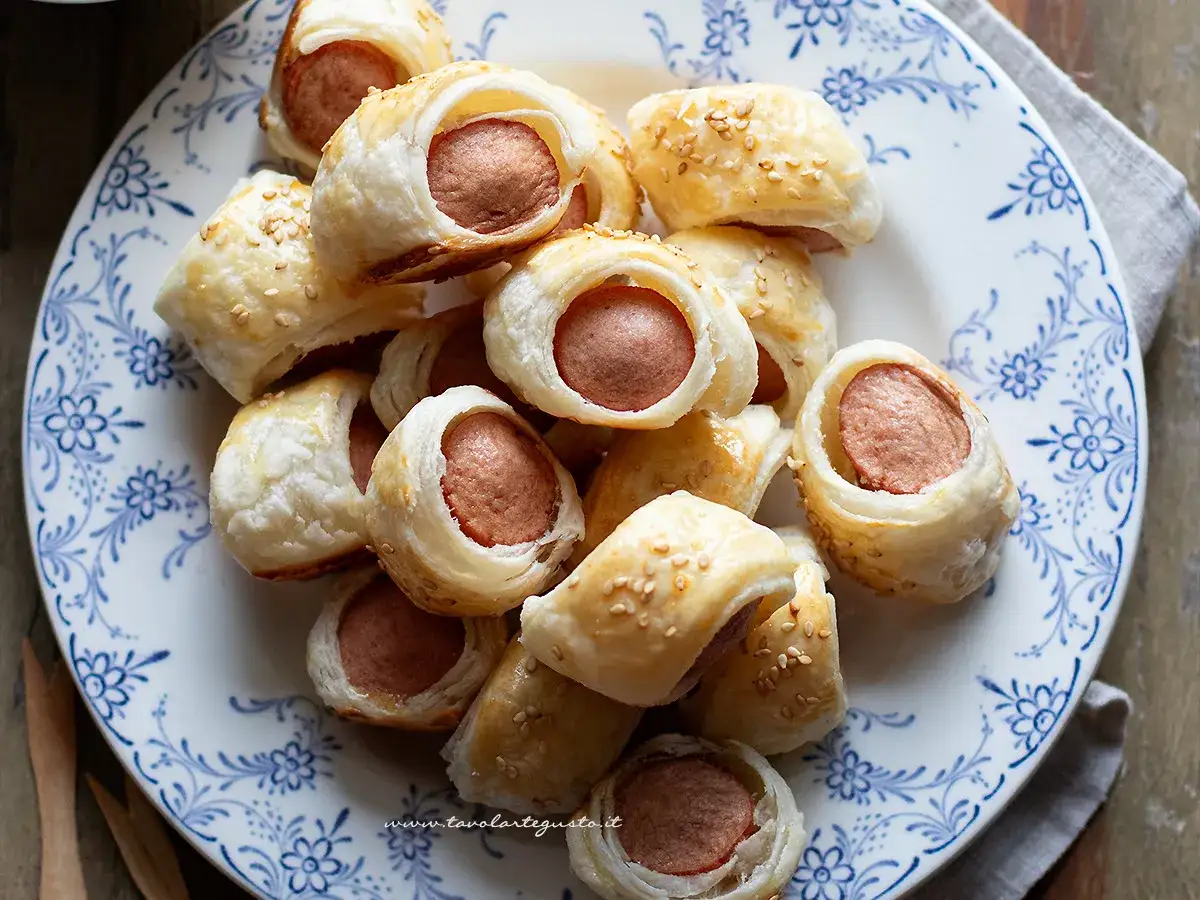 Rustici con wurstel dorati e croccanti, cosparsi di semi di sesamo, serviti su un piatto bianco con decori blu.