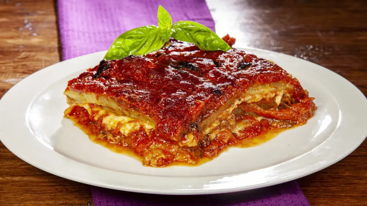 Una fetta di parmigiana di melanzane, ricca e saporita, guarnita con basilico fresco, su un piatto bianco.