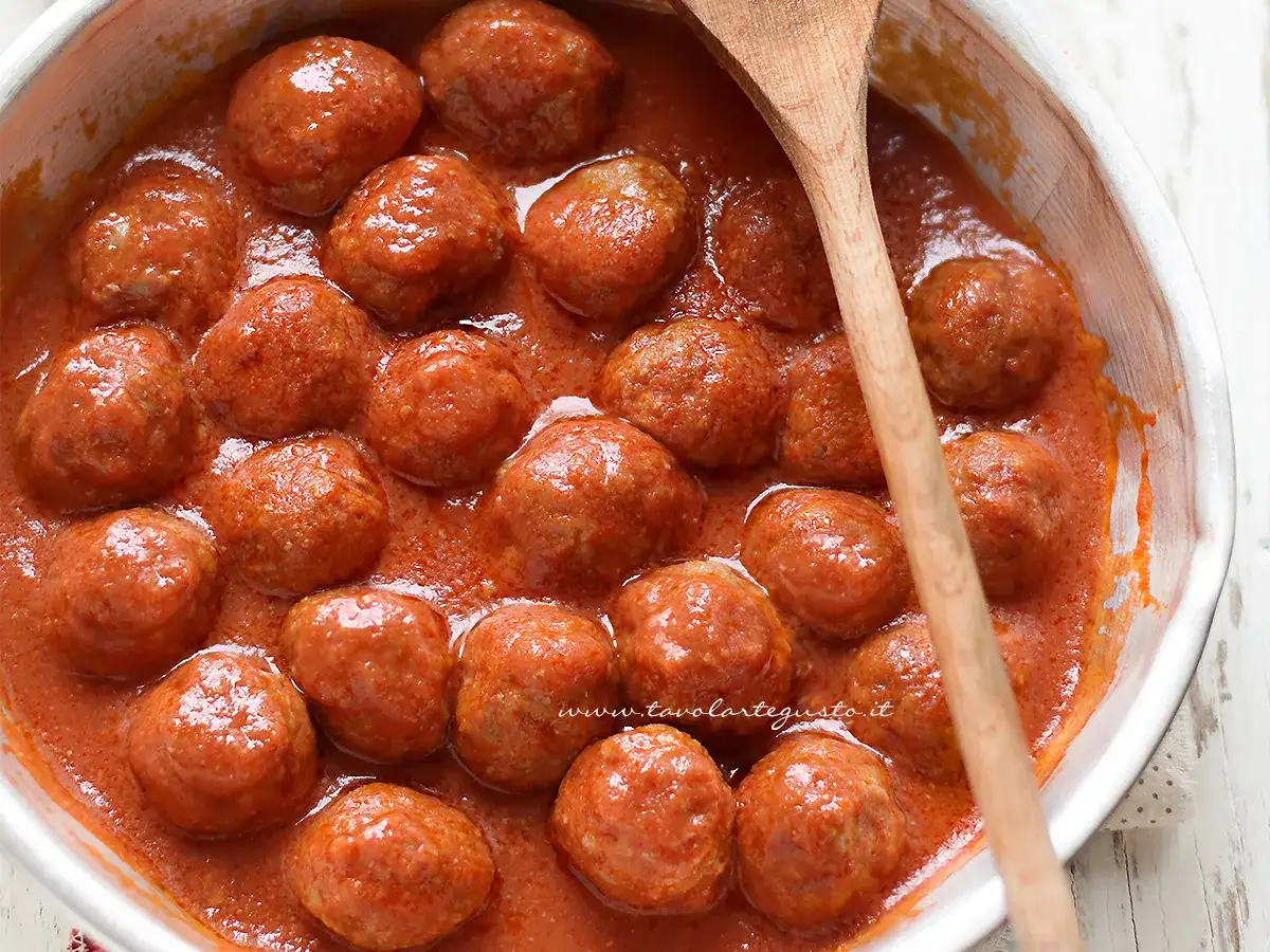 Polpette di carne succulente in un sugo rosso, pronte per essere gustate. Un cucchiaio di legno attende di servirle.