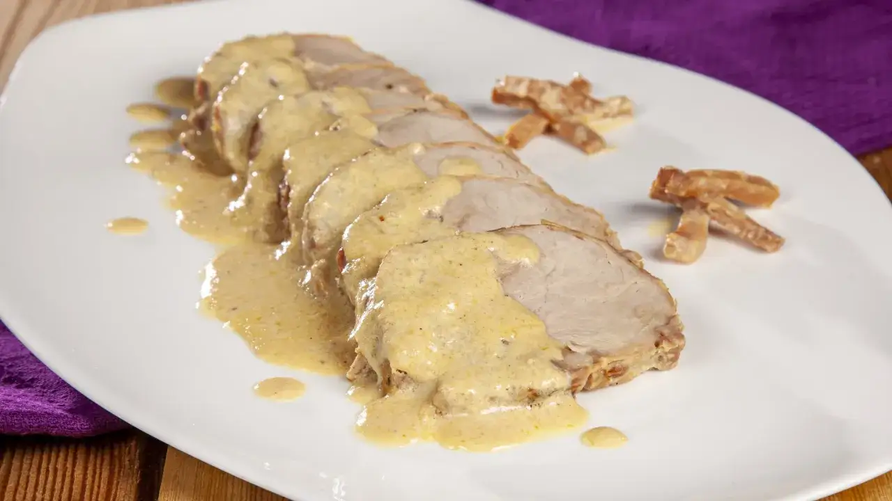 Arista al latte affettata su un piatto bianco, ricoperta da una cremosa salsa. Accompagnata da bastoncini croccanti.