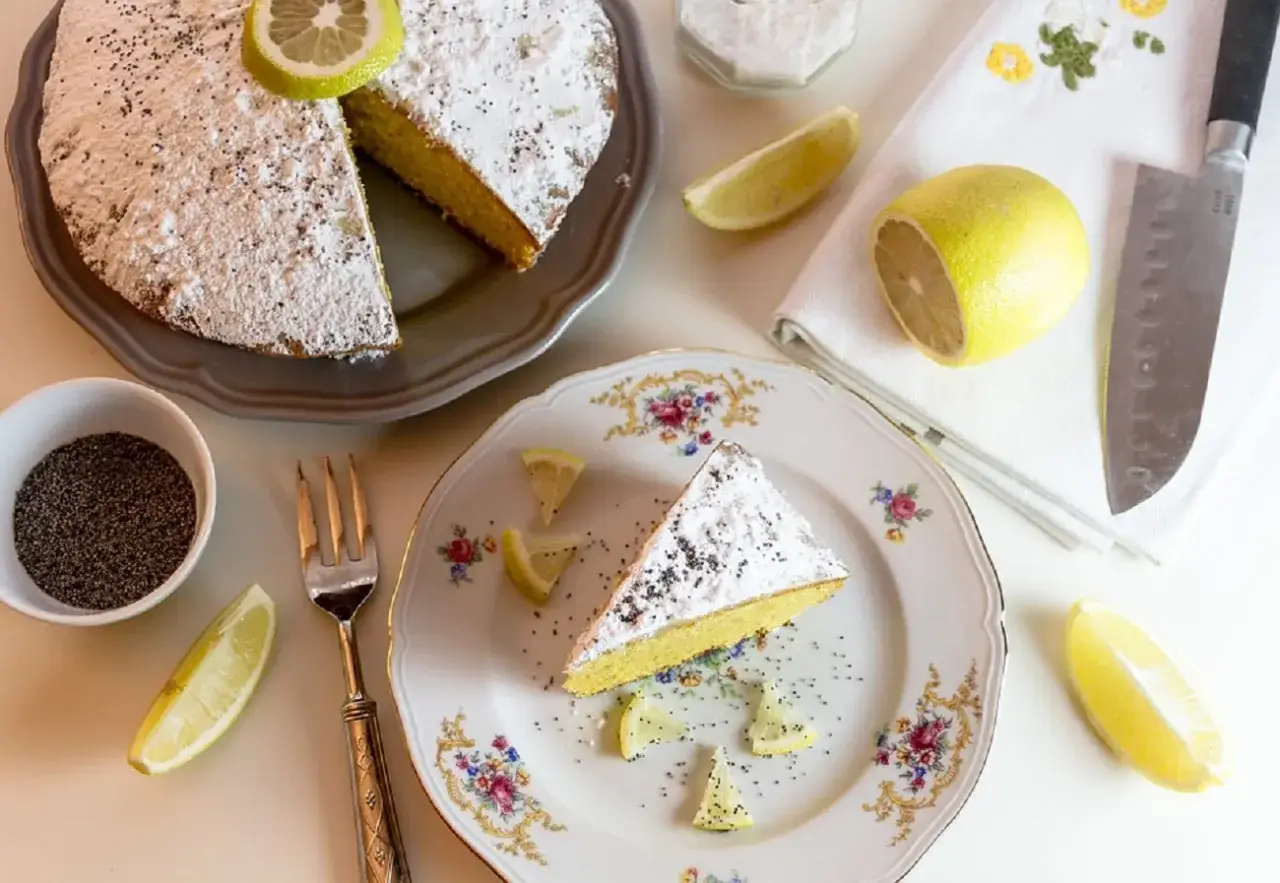 Un delizioso plumcake al limone, spolverato di zucchero a velo e semi di papavero, con spicchi di limone fresco e una forchetta d'argento.