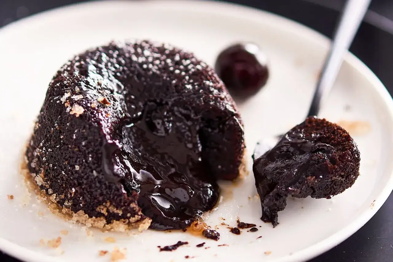 Tortino al cioccolato con cuore fuso, cosparso di zucchero a velo, accanto a una ciliegia scura.