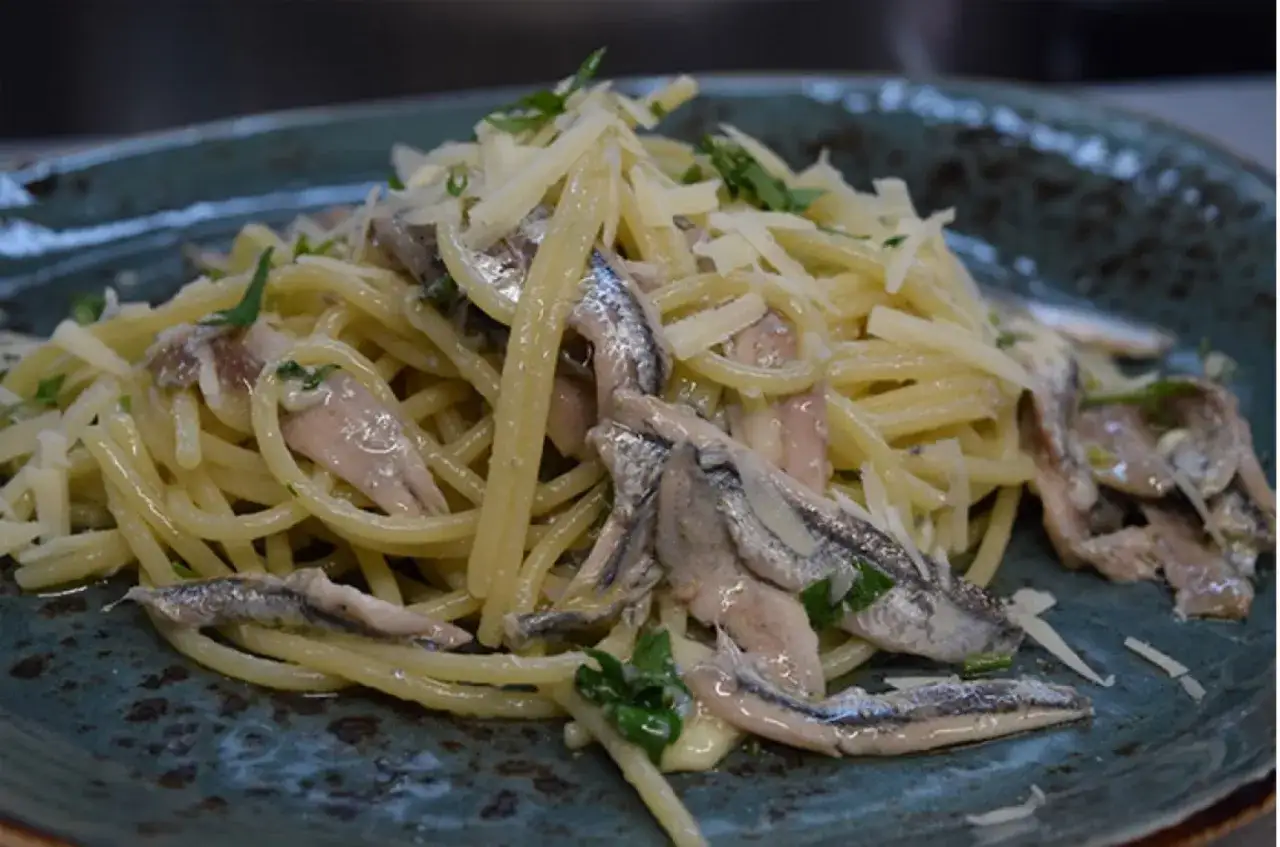 Spaghetti conditi con alici fresche, formaggio grattugiato e prezzemolo su un piatto blu.