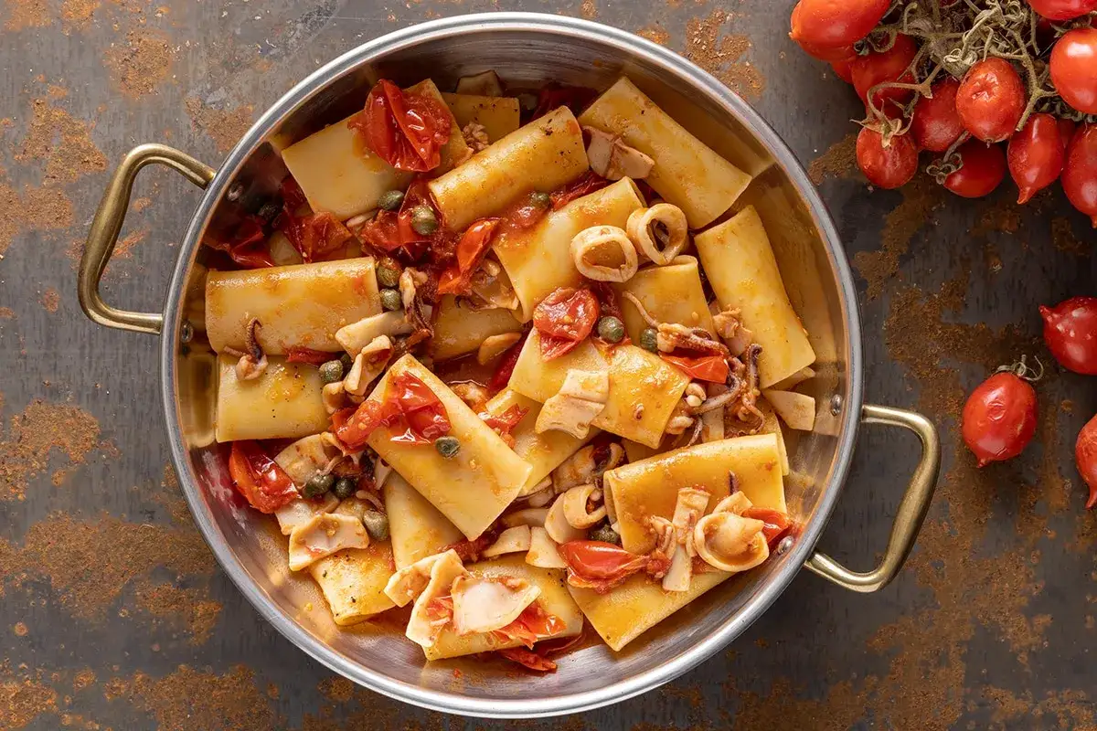 Paccheri con calamari, pomodorini e capperi in un tegame di metallo. Un piatto invitante che profuma di mare.