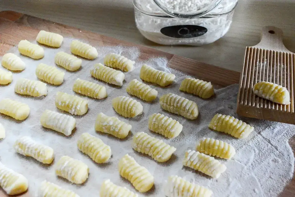 Gnocchi di patate ricetta originale pronti per essere cotti. Un vassoio pieno di morbidi gnocchi fatti in casa, spolverati di farina, con un rigagnocchi in legno sullo sfondo.
