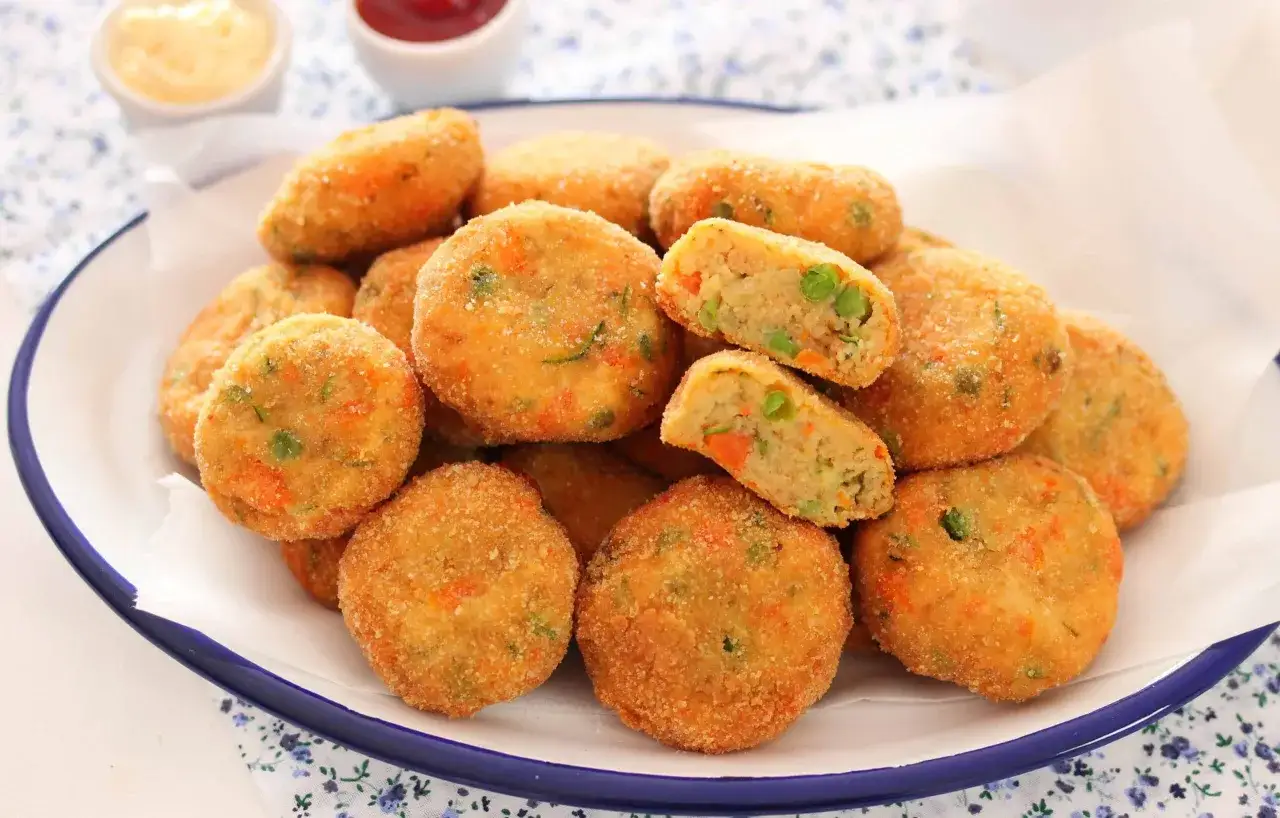 Piatto colmo di polpette di verdure dorate e croccanti, con piselli e carote visibili all'interno. Due ciotoline con salse sullo sfondo.