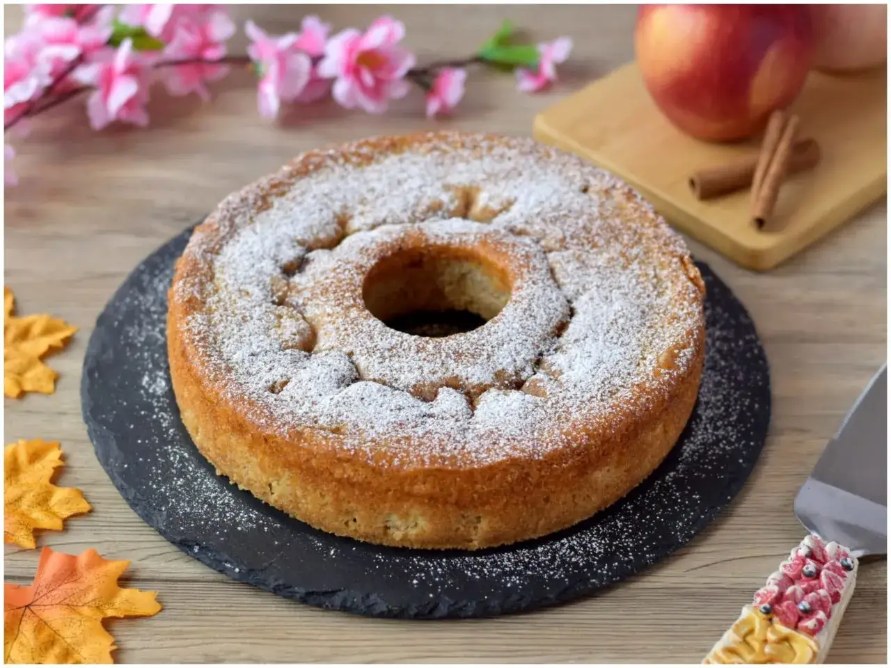 Deliziosa ciambella alle mele, cosparsa di zucchero a velo, su un piatto di ardesia. Sullo sfondo, mele rosse e cannella.