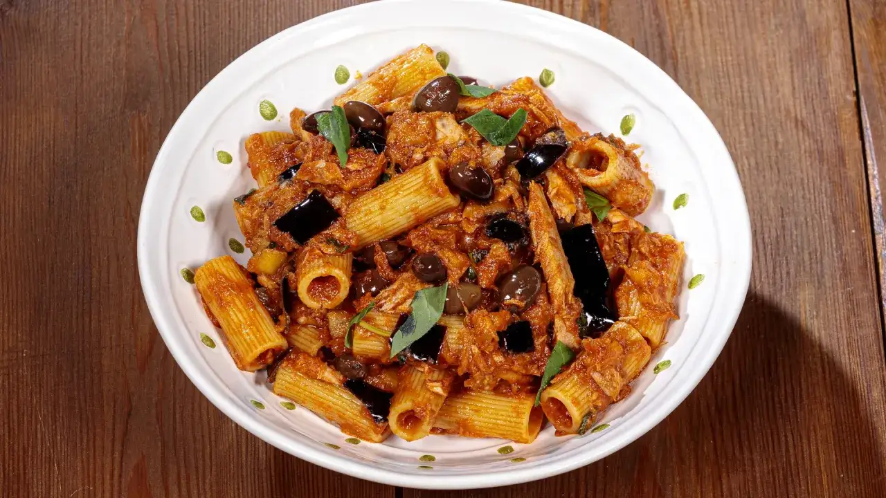Piatto di pasta con melanzane, tonno e olive nere, guarnito con foglie di basilico fresco.