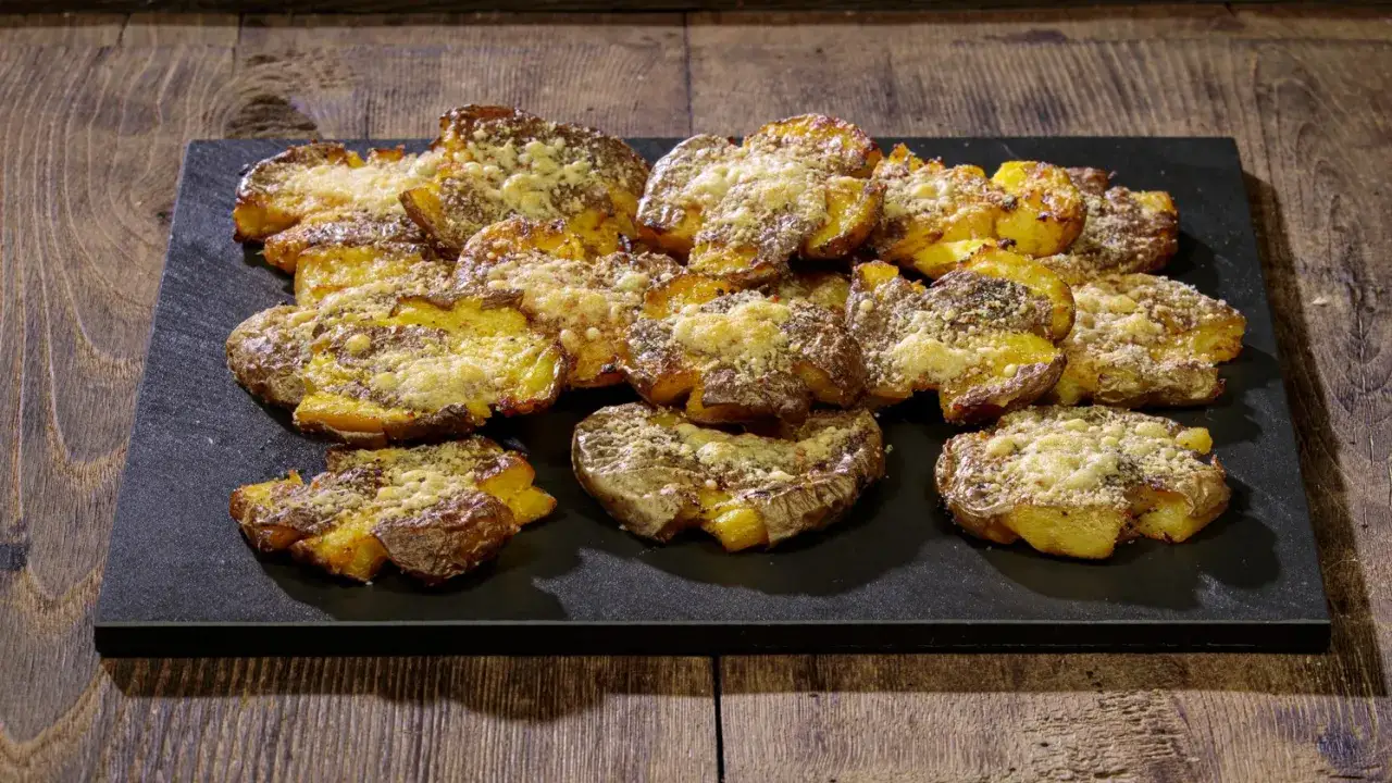 Patate schiacciate al forno, dorate e croccanti, cosparse di formaggio grattugiato e erbe aromatiche, servite su una pietra scura.