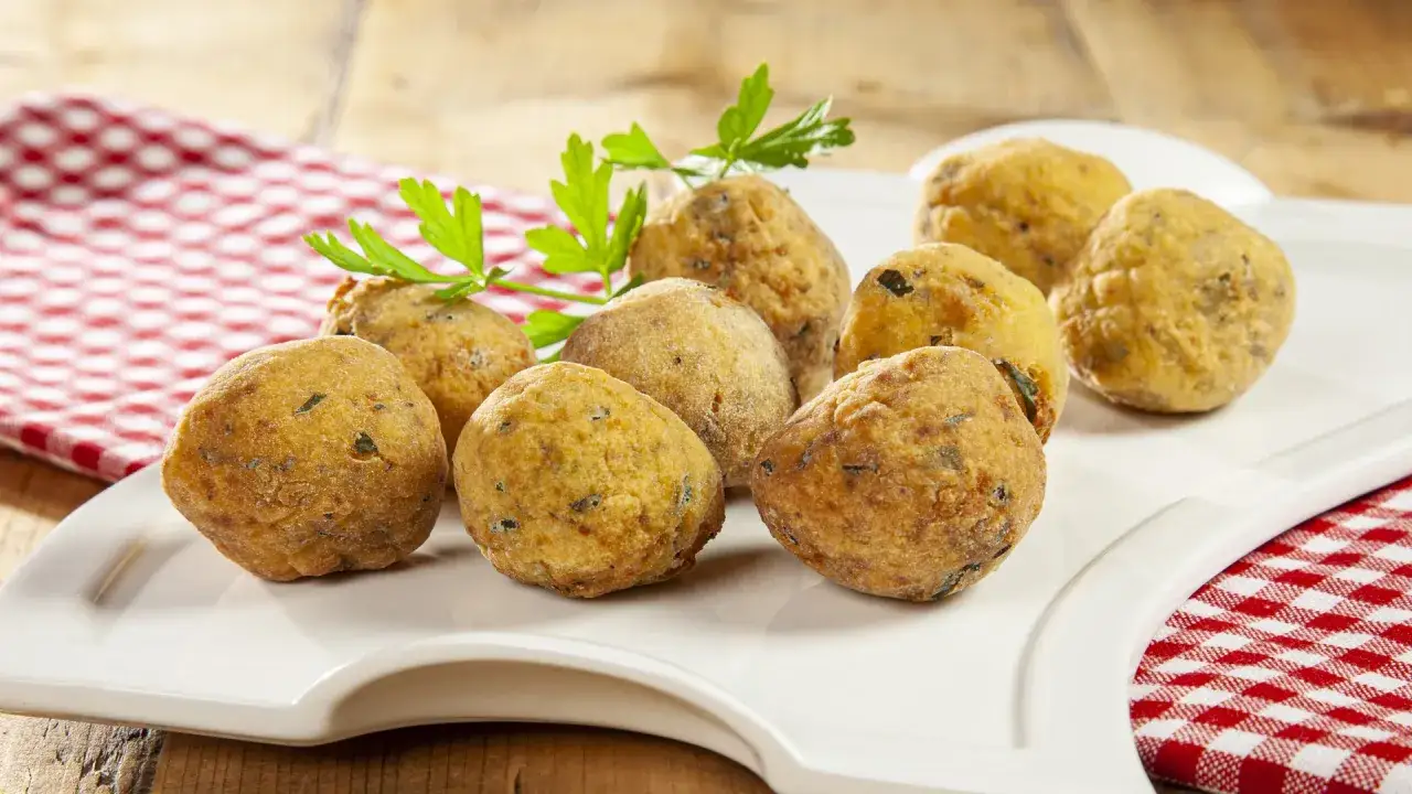 Un piatto bianco con polpette di pane dorate e croccanti, guarnite con prezzemolo fresco. Un tovagliolo a quadri rossi e bianchi completa la scena.
