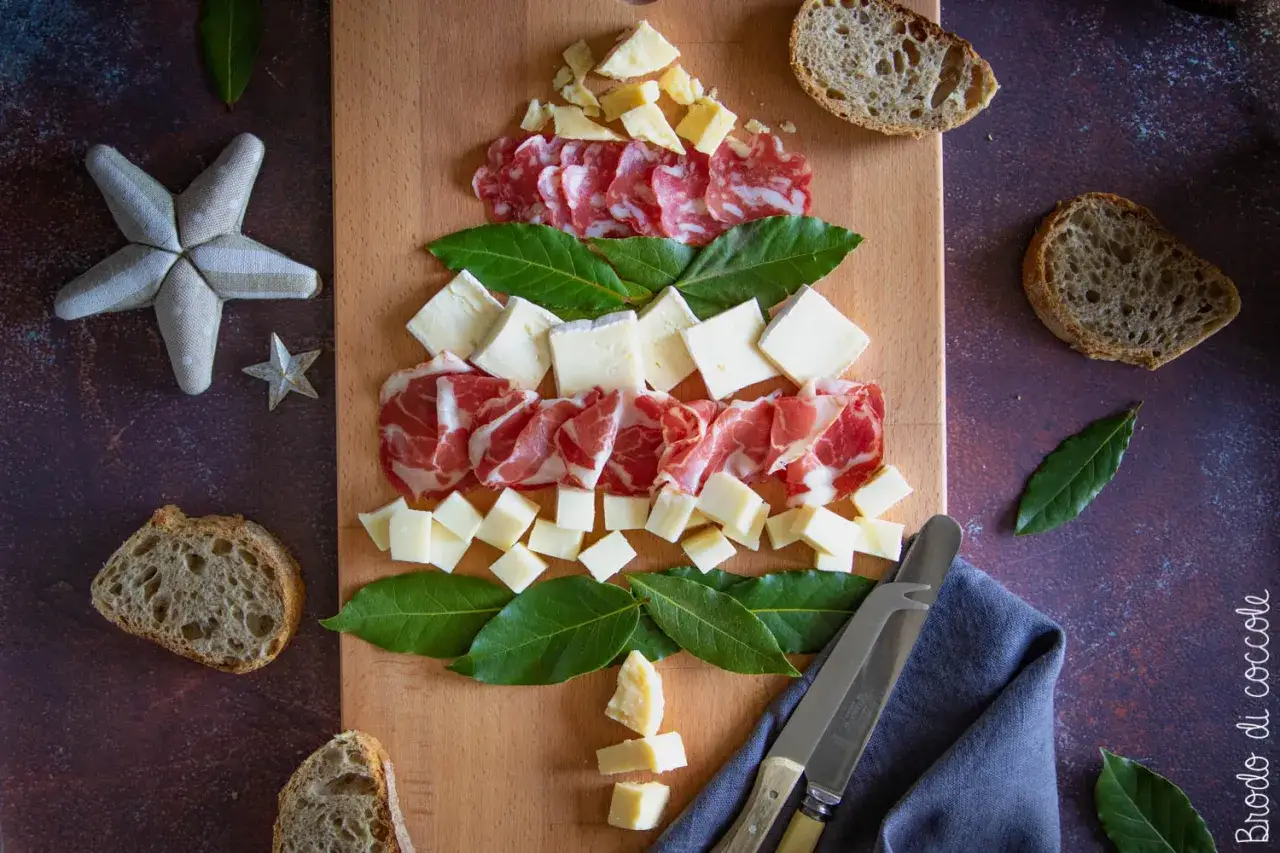 Un albero di Natale fatto con un tagliere di salumi e formaggi, decorato con foglie di alloro e pane rustico.