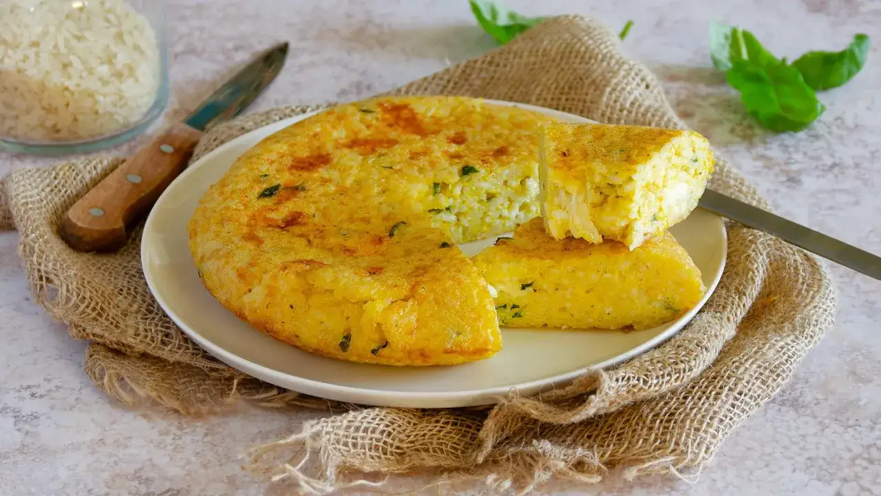 Una dorata frittata di riso, con erbe aromatiche, servita su un piatto bianco, accanto a un barattolo di riso e foglie di basilico.