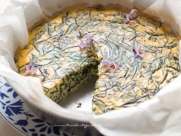 Frittata di agretti appena sfornata, decorata con fiori viola, in una teglia foderata di carta forno.