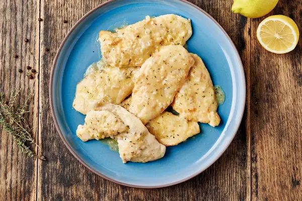 Piatto di petto di pollo al limone, condito con erbe aromatiche e servito su un piatto blu. Accanto, limone fresco e pepe nero.