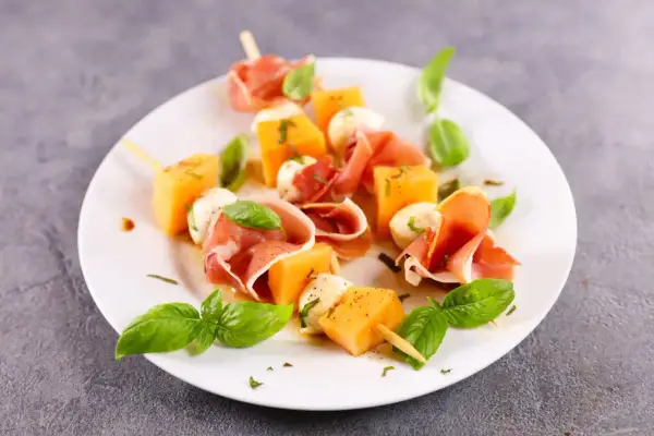 Spiedini di prosciutto e melone con mozzarelline e basilico fresco, un antipasto estivo perfetto.