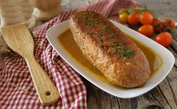 Polpettone dorato in padella, guarnito con erba cipollina, su un piatto bianco con salsa, accanto a pomodorini e una spatola di legno.