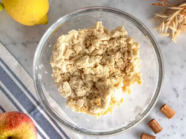 Impasto friabile per crumble di mele in una ciotola di vetro, con limone, mela e cannella sullo sfondo.